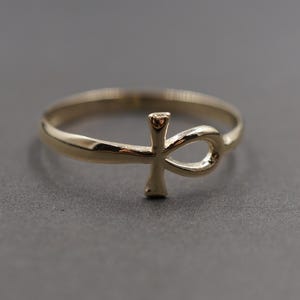 Puede incluir: Un anillo dorado con un símbolo Ankh. El anillo tiene una banda delgada y el Ankh se muestra prominentemente en el centro. El anillo está sobre un fondo gris, resaltando su diseño.