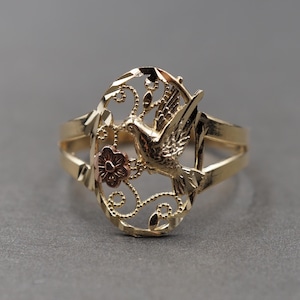 Puede incluir: Un anillo de oro con un diseño ovalado de filigrana, un motivo central de ave y un pequeño acento de flor en oro rosa. La banda está pulida y el marco ovalado tiene un borde texturizado y facetado, creando una apariencia detallada y compleja.