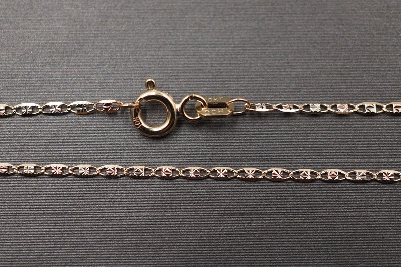 May include: A gold chain necklace with a delicate, patterned design. The chain is made of small, oval-shaped links with a diamond-shaped pattern on each link. The chain has a lobster clasp closure.