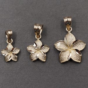 May include: Three gold-colored flower pendants of varying sizes are displayed against a dark gray background. Each pendant features a five-petal floral design with textured detailing and a loop for a chain.