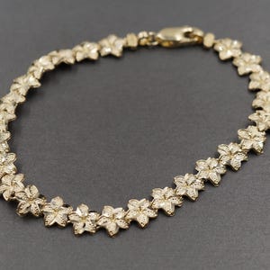 May include: A gold-colored bracelet featuring a floral design. The bracelet is composed of multiple flower-shaped links connected together. The clasp is also gold-colored. The bracelet is displayed on a dark gray surface.