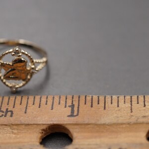 May include: A gold-toned ring featuring a central circular motif with a cherub figure inside, flanked by small clear stones. The ring is positioned next to a wooden ruler showing a scale in inches to provide a clear sense of its physical size.