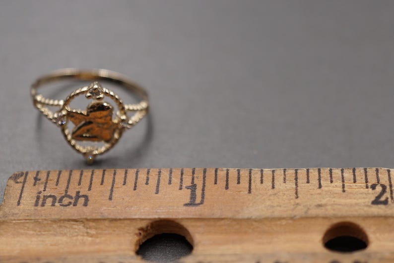 May include: A gold-toned ring featuring a central circular motif with a cherub figure inside, flanked by small clear stones. The ring is positioned next to a wooden ruler showing a scale in inches to provide a clear sense of its physical size.