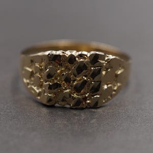 May include: A gold nugget ring with a textured surface. The ring features a wide band and a rounded top, displaying a detailed, irregular pattern. The ring is likely made of gold or a gold-toned metal.