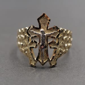 May include: A gold ring featuring a central crucifix design with a silver-toned figure of Jesus. The band has a textured, nugget-style finish and includes two additional smaller cross motifs on either side of the central crucifix element.