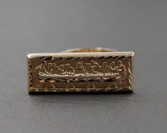 May include: A rectangular gold ring featuring a detailed relief carving of The Last Supper on its face. The surface has a polished finish with textured, diagonal line detailing along the border, creating a distinct contrast against the scene.