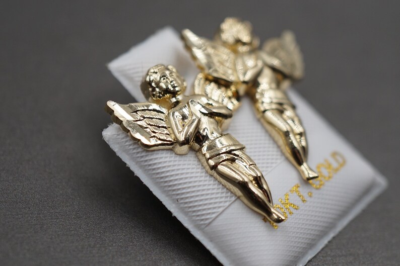 May include: A pair of gold angel earrings. The angels are depicted with wings and are facing each other. The earrings are on a white background with the text "14K GOLD" printed on it.
