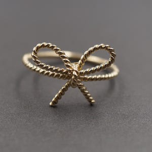 May include: A gold-coloured ring with a bow design. The bow and ring band have a twisted rope texture. The ring is set against a dark grey background, highlighting its delicate details. A piece of jewellery.