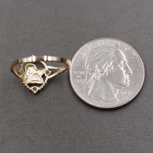 May include: A gold-toned ring featuring a central cherub or angel motif with a scalloped, openwork band design. The ring is positioned next to a United States quarter coin for scale, which serves as a reference for the size of the jewelry piece.