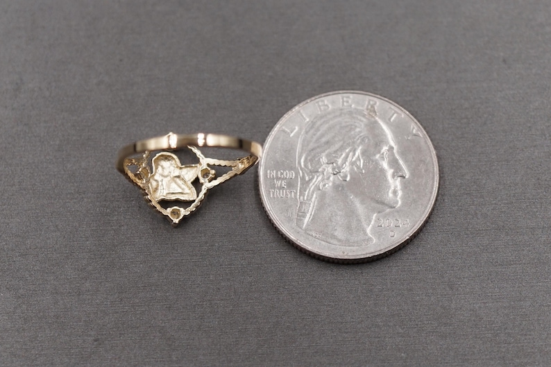 May include: A gold-toned ring featuring a central cherub or angel motif with a scalloped, openwork band design. The ring is positioned next to a United States quarter coin for scale, which serves as a reference for the size of the jewelry piece.