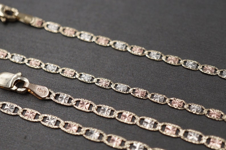 May include: Three gold chains with a tri-color design. The chains are made of yellow, white, and rose gold. The chains are linked together with a square-shaped link. The chains are on a black surface.