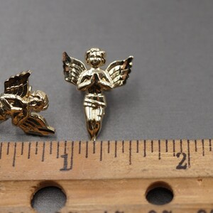 May include: Two gold-colored angel figurines with wings, one with hands clasped in prayer and the other with wings spread out. The figurines are about 1 inch tall and are shown next to a ruler.
