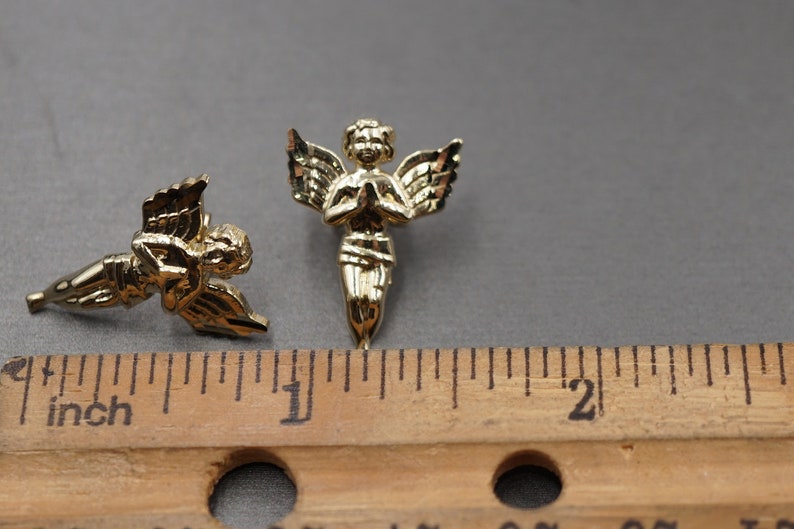 May include: Two gold-colored angel figurines with wings, one with hands clasped in prayer and the other with wings spread out. The figurines are about 1 inch tall and are shown next to a ruler.