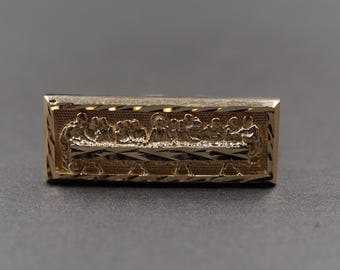May include: A rectangular gold-toned metal bar featuring a detailed embossed relief of The Last Supper. The scene depicts figures seated at a long table with a textured background and a diagonal striped border framing the central imagery.