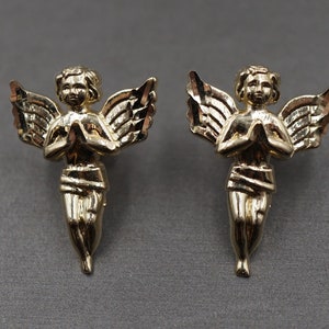 May include: A pair of gold angel earrings with wings and hands clasped in prayer.