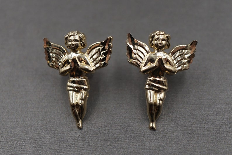 May include: A pair of gold angel earrings with wings and hands clasped in prayer.