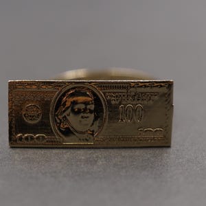 Op de afbeelding: Een goudkleurige rechthoekige ring met een gedetailleerde afbeelding van een biljet van $100. Het ontwerp bevat het portret van Benjamin Franklin en het getal "100". De ring is een uniek accessoire.