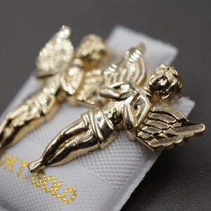 May include: Two gold-tone angel figurines with wings outstretched. The figurines are on a white background with the word "Gold" printed in gold lettering.