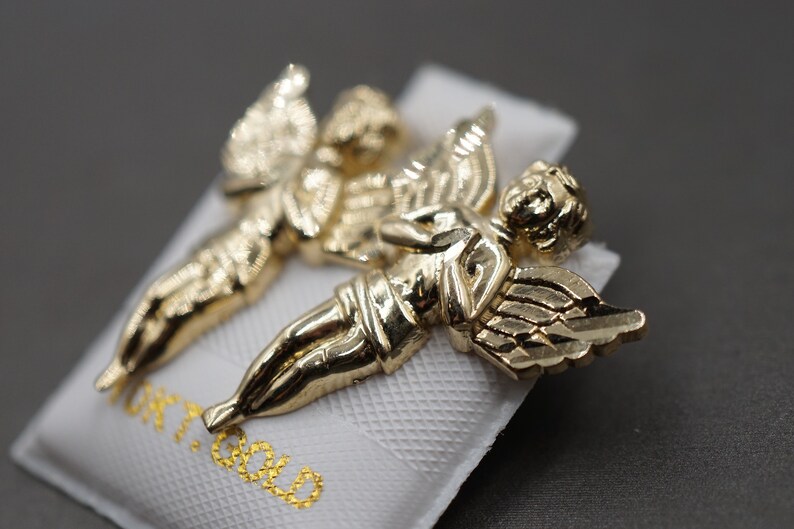 May include: Two gold-tone angel figurines with wings outstretched. The figurines are on a white background with the word "Gold" printed in gold lettering.