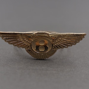 May include: A gold-coloured emblem featuring a winged design with a central circular element. The emblem has a detailed, embossed appearance, with the letter "B" visible in the centre. The wings extend outwards on either side.