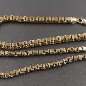 Könnte beinhalten: Zwei goldfarbene Armbänder mit einem strukturierten, gewebten Design. Jedes Armband hat einen Verschluss. Die Armbänder werden auf einer grauen Oberfläche präsentiert. Die Armbänder sind wahrscheinlich aus Metall.