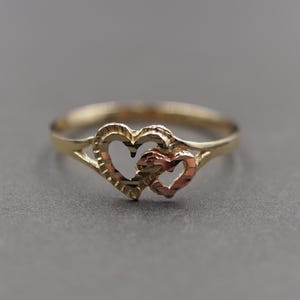 Puede incluir: Un anillo de oro con dos formas de corazón entrelazadas. Un corazón es dorado y el otro es una mezcla de oro y oro rosa. El anillo está sobre un fondo gris.