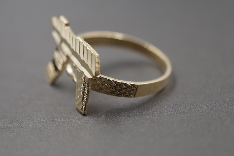May include: A gold-colored ring with a detailed design of a gun. The ring is made of metal and has a textured finish.