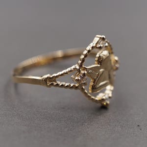 May include: A gold ring featuring a central angel figure within a circular frame. The band and the circular frame have a twisted rope texture. Small, clear, round stones are set at the top, bottom, and sides of the circular frame surrounding the angel.