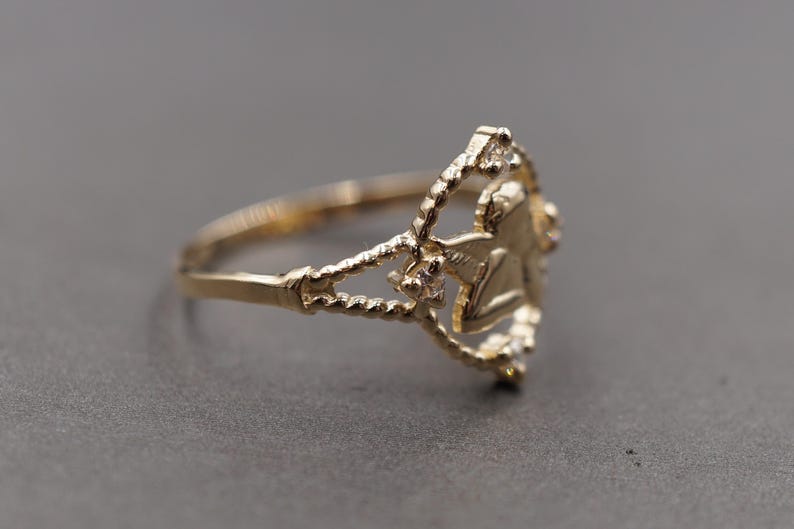 May include: A gold ring featuring a central angel figure within a circular frame. The band and the circular frame have a twisted rope texture. Small, clear, round stones are set at the top, bottom, and sides of the circular frame surrounding the angel.