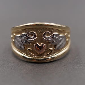 May include: A gold ring with two silver elephants and a rose gold heart. The ring has a textured band and a polished finish. The design is symmetrical and detailed, set against a grey background. The ring is a statement piece.