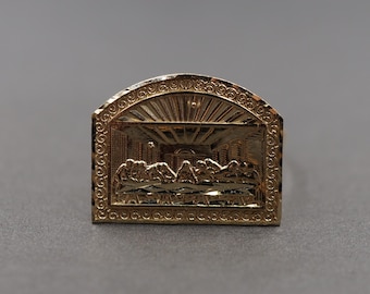 May include: A gold-colored ring with a raised image of the Last Supper. The ring is detailed with a textured border and a sunburst design.