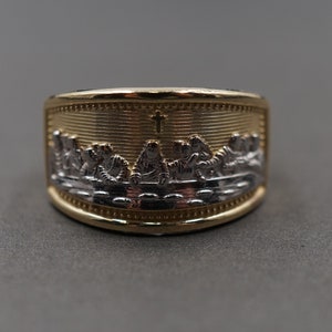 Pode incluir: Um anel de ouro e prata com uma representação da Última Ceia e uma cruz. O anel é feito de metal de dois tons e tem um design detalhado.
