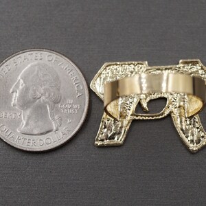 May include: Gold-tone ring featuring a detailed design of a handgun. The ring is shown next to a US quarter for size comparison.