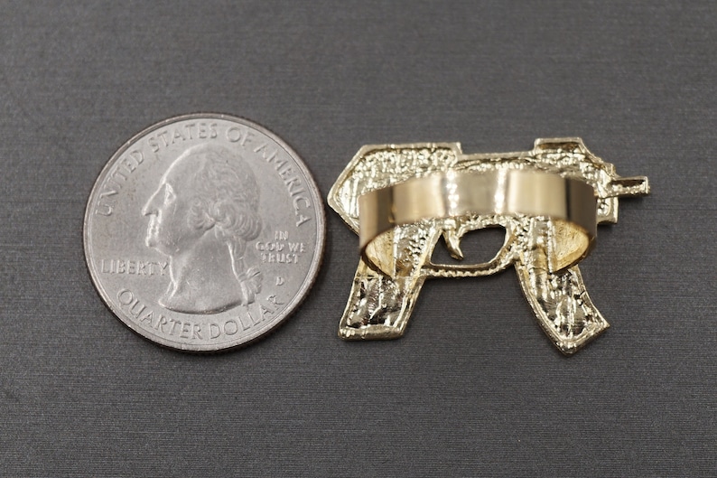 May include: Gold-tone ring featuring a detailed design of a handgun. The ring is shown next to a US quarter for size comparison.