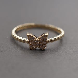 May include: A gold-colored ring with a butterfly design. The butterfly is encrusted with small, clear stones. The band has a beaded texture. The ring is set against a gray background.
