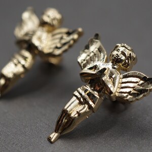 May include: A pair of gold-tone angel earrings. Each earring features a detailed angel with wings and a delicate, feminine design.