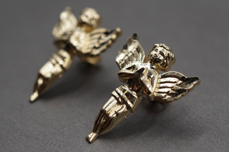 May include: A pair of gold-tone angel earrings. Each earring features a detailed angel with wings and a delicate, feminine design.
