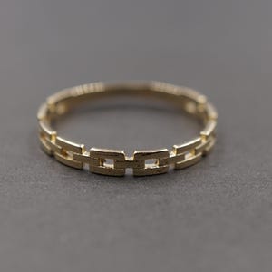 Könnte beinhalten: Ein goldfarbener Ring mit einem durchgehenden Kettenglied-Design. Das Band besteht aus kleinen, rechteckigen Metallsegmenten, die miteinander verbunden sind und ein sich wiederholendes Muster um den gesamten Umfang bilden.