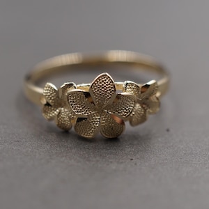 May include: A gold-colored ring featuring a floral design. The ring has a band and a cluster of three-dimensional flowers. The flowers have detailed textures, and the ring is set against a gray background. The ring is a piece of jewelry.