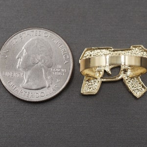 May include: Gold-tone ring featuring a detailed design of a handgun. The ring is shown next to a US quarter for size comparison.