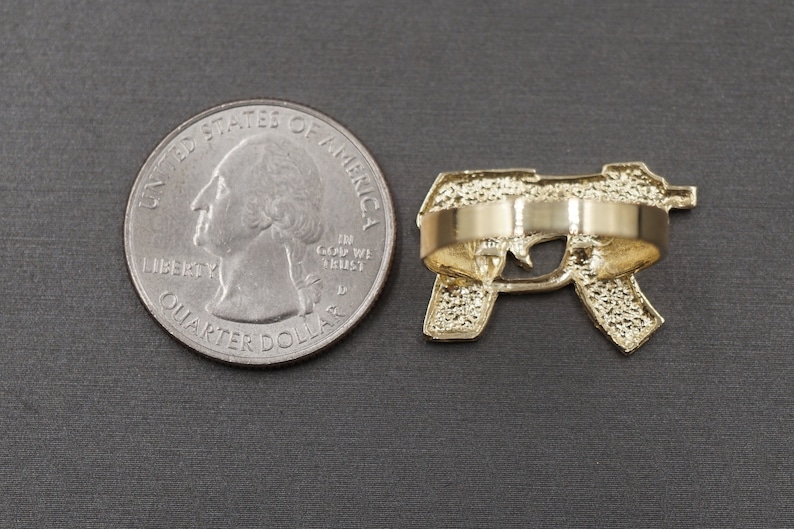May include: Gold-tone ring featuring a detailed design of a handgun. The ring is shown next to a US quarter for size comparison.