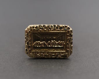 May include: A gold-colored ring with a rectangular face. The face features a detailed relief of the Last Supper scene, framed by a textured, nugget-like border. The ring is likely made of gold or a gold-toned metal.