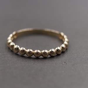 May include: A gold ring with a beaded design. The ring features a series of small, evenly spaced spheres around the band. The ring is set against a dark gray background.