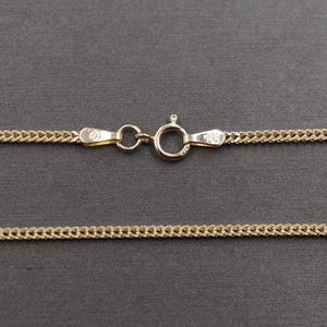 Op de afbeelding: Een goudkleurige ketting met een karabijnsluiting. De ketting heeft een curb-link ontwerp. De sluiting en ketting worden getoond tegen een donkergrijze achtergrond.