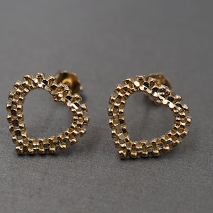 May include: A pair of gold-coloured stud earrings, each in the shape of a heart. The heart shape is open in the middle and outlined with small, textured details. The earrings are shown on a grey surface.