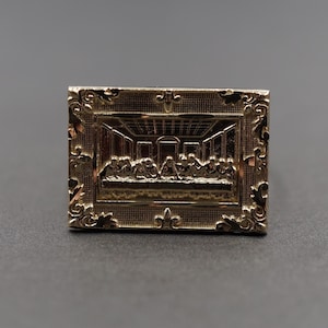 Può includere: Un anello color oro con una cornice ornata che raffigura l'Ultima Cena. L'anello presenta dettagli intricati e un design in rilievo.