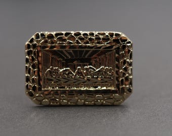 May include: A gold-colored ring with a rectangular face. The face features a detailed relief of the Last Supper scene. The ring's edges have a textured, pebble-like design, adding visual interest to the jewelry.