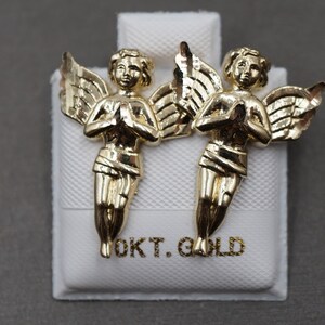 May include: A pair of gold angel earrings with wings outstretched and hands clasped in prayer. The earrings are displayed on a white box with the text "OKT. GOLD" printed on it.