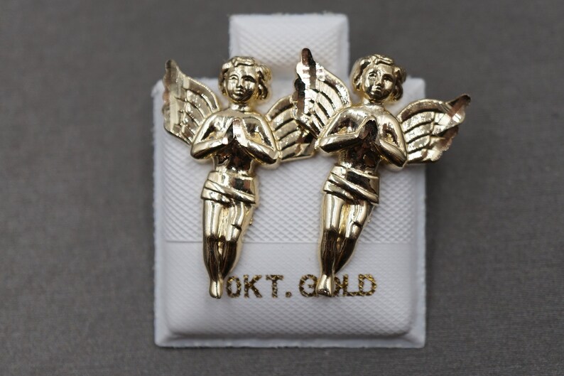 May include: A pair of gold angel earrings with wings outstretched and hands clasped in prayer. The earrings are displayed on a white box with the text "OKT. GOLD" printed on it.