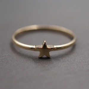 Puede incluir: Un anillo dorado con un diseño en forma de estrella. El anillo presenta una banda delicada y una pequeña estrella en relieve en el centro. El anillo probablemente está hecho de oro o metal dorado.